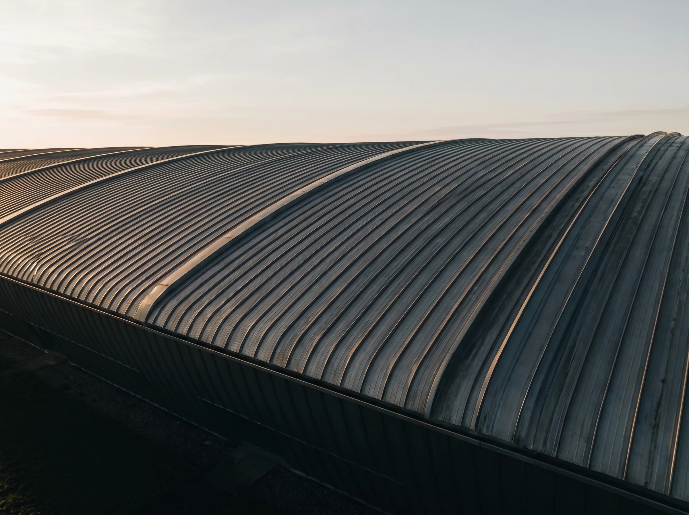 Drone aerial of a curved standing-seam aluminium roof at golden hour, brass-warm light catching the seam ribs